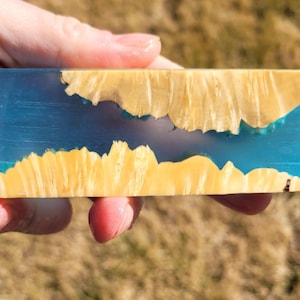 Könnte beinhalten: Ein rechteckiger Block aus klarem Harz mit einem leuchtenden Blauton, der organische, hellgelbe Holzeinschlüsse aufweist. Das Holz scheint in das Harz eingebettet zu sein und ein einzigartiges, natürliches Design zu erzeugen. Der Block wird in einer Hand gehalten.