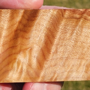 May include: A close-up of a piece of wood with a unique grain pattern. The wood is a light brown color with streaks of darker brown and red. The grain is swirling and wavy, creating a beautiful and interesting pattern.