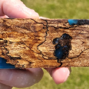 Könnte beinhalten: Ein rechteckiger Block aus stabilisiertem Holz mit einem komplexen, wirbelnden Maserungsmuster in Braun- und Beigetönen, akzentuiert durch dunkle, komplizierte spinnennetzartige Linien und eingebettete Abschnitte aus durchscheinendem, blauem Harz.
