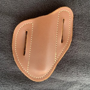 May include: A brown leather holster with two vertical slots and white stitching. The holster has a curved shape and is designed to hold a small object. The leather has a smooth texture.