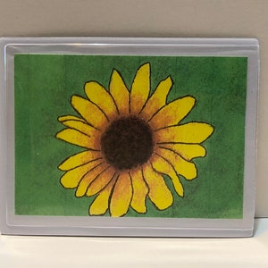 May include: A yellow sunflower with a brown center is drawn on a green background. The image is framed in a white border.