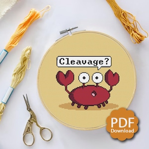 May include: A cross-stitch pattern featuring a red crab with large eyes and a speech bubble that says "Cleavage?" The pattern is on a yellow background and is framed in a wooden hoop.