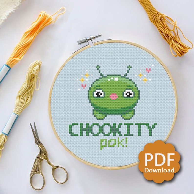 Puede incluir: Un patr&oacute;n de punto de cruz con un extraterrestre verde con una expresi&oacute;n linda, rodeado de corazones y estrellas. El texto "Chookitty Pok!" est&aacute; escrito debajo del extraterrestre.