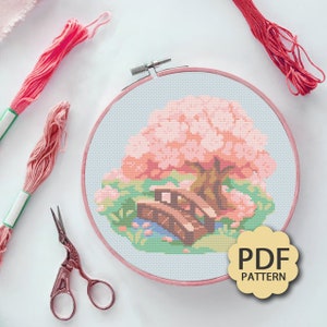 May include: A cross-stitch pattern featuring a pink cherry blossom tree with a wooden bridge in a green garden. The pattern is displayed in a round embroidery hoop with a light blue background. The text "PDF PATTERN" is visible in the lower right corner of the image.