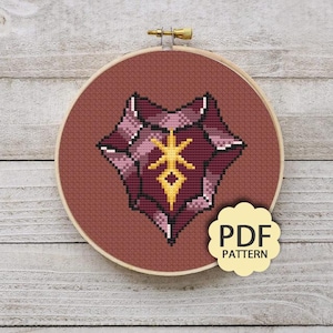 May include: Cross-stitch embroidery in a wooden hoop. The design features a geometric shape in shades of burgundy, pink, and white, with a gold-colored central emblem. A small, cream-colored tag with the text "PDF PATTERN" is attached.