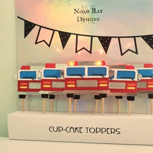 May include: Set of 6 red and silver fire truck cupcake toppers with black wheels and yellow headlights. The toppers are on wooden sticks and are displayed on a white stand with the text "CUP-CAKE TOPPERS".