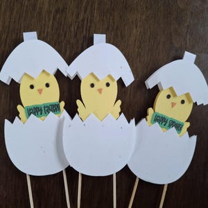 May include: Three yellow chick paper cutouts with green banners saying "Happy Easter" and "Happy Spring" are inside white egg-shaped paper cutouts. The cutouts are on wooden sticks.