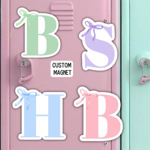 May include: Four colorful magnetic letters with bows, spelling "BHSB", on a pink locker door. The letters are green, purple, light blue, and pink. The letters are decorated with bows in matching colors. The locker door has a silver handle and a silver lock.