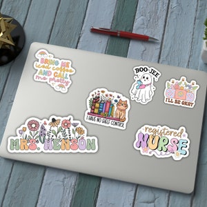 K&ouml;nnte beinhalten: Eine Sammlung bunter Aufkleber auf einem Laptop. Die Aufkleber zeigen verschiedene Designs, darunter ein Geist mit einem Getr&auml;nk, eine Katze mit B&uuml;chern, Blumenarrangements und die S&auml;tze "Bring me iced coffee and call me pretty", "I have no shelf control" und "Registered Nurse."