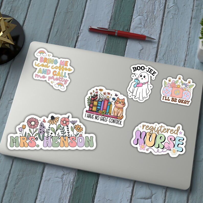 K&ouml;nnte beinhalten: Eine Sammlung bunter Aufkleber auf einem Laptop. Die Aufkleber zeigen verschiedene Designs, darunter ein Geist mit einem Getr&auml;nk, eine Katze mit B&uuml;chern, Blumenarrangements und die S&auml;tze "Bring me iced coffee and call me pretty", "I have no shelf control" und "Registered Nurse."