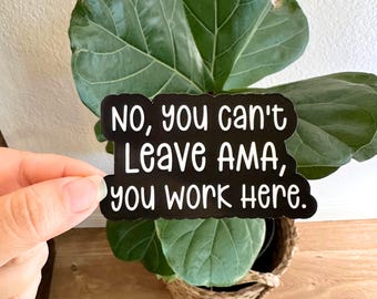 May include: A black sticker with white text that reads "NO, you can't Leave AMA, you work HERE." The sticker is held in front of a green plant with large leaves in a woven basket.
