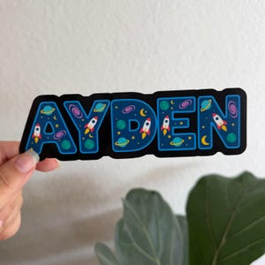 May include: A black sticker with the name "AYDEN" in blue bubble letters. Each letter is decorated with a space theme, including rockets, planets, stars, and galaxies. The sticker is held by a hand.