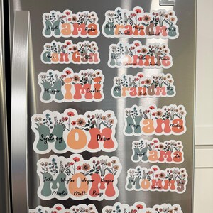 May include: A collection of floral-themed refrigerator magnets. Each magnet features a name, such as "Mama" or "Grandma", in a stylized font, with a vase of flowers. The color palette includes orange, green, and white.