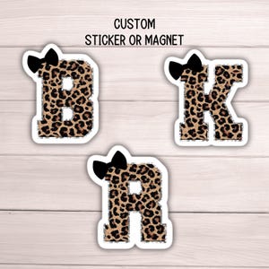 May include: Three custom stickers or magnets featuring the letters B, K, and R. Each letter has a leopard print pattern and a black bow at the top. The text "CUSTOM STICKER OR MAGNET" is at the top.