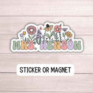 K&ouml;nnte beinhalten: Ein Aufkleber oder Magnet mit dem Namen "MRS. HENSON" in bunten, abgerundeten Buchstaben, umgeben von einem Blumendesign mit rosa, orangefarbenen, gelben und lila Blumen. Der Text "STICKER OR MAGNET" steht darunter.