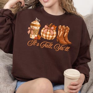 May include: A dark brown crewneck sweatshirt featuring a graphic print of a pumpkin spice coffee cup, a plaid pumpkin with a bow, and a pair of brown cowboy boots. The text "It's Fall, Ya'll" is printed in a cursive orange font below the graphics.