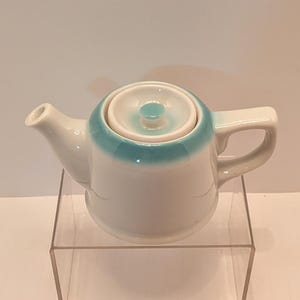 May include: A small, white ceramic teapot with a turquoise blue band around the top and lid. The teapot has a curved handle and a spout. The lid has a small knob. The teapot is displayed on a clear acrylic stand.