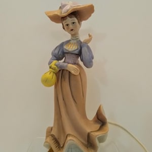May include: A porcelain figurine of a woman in a long dress and hat. The figurine is painted in muted tones of peach, lavender, and yellow. The woman is holding a yellow purse. The figurine is on a clear acrylic stand.