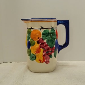 May include: Cream-colored ceramic pitcher with a blue handle and rim. The pitcher is decorated with colorful fruit including oranges, grapes, and pears. The fruit is painted in bright colors with green leaves. The pitcher is a decorative item.