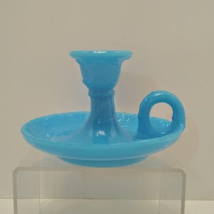 May include: A light blue glass candlestick holder with a round base and a handle. The candlestick has a detailed floral design around the base and the candle cup. The handle is a loop shape.