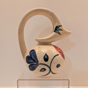 May include: Cream-colored ceramic pitcher with a curved handle and floral designs. The pitcher features blue and red flowers with green leaves. The handle curves over the top, ending in a pointed spout. A decorative piece for the home.