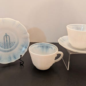 May include: A set of vintage white teacups and saucers with a blue rim and a decorative plate with a blue design. The teacups and saucers are displayed on a black surface and a clear stand.