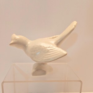 May include: A cream-colored ceramic bird figurine with a detailed wing design and a raised crest. The bird is perched on a small base, showcasing a smooth, glossy finish. The figurine is displayed on a clear acrylic surface.
