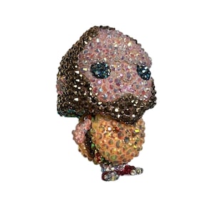 Könnte beinhalten: Eine mit Kristallen besetzte Figur eines Charakters mit großem Kopf und kleinem Körper. Die Figur ist mit schillernden Kristallen in Rosa-, Braun- und Goldtönen bedeckt. Die Augen sind dunkelblau und die Füße rot.