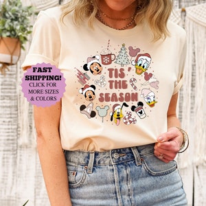 May include: A beige t-shirt with a festive Christmas design featuring Mickey Mouse, Minnie Mouse, Donald Duck, and other Disney characters. The text on the shirt reads "Tis' the Season".
