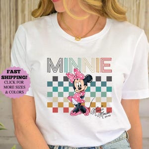 以下が含まれることがあります： ミニーマウスのグラフィックが特徴の白いTシャツ。デザインには、マルチカラーのフォントで書かれた「MINNIE」の文字、市松模様、ピンクのドレスを着たミニーマウスの漫画の画像が含まれています。