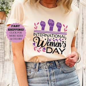 May include: Sand-colored t-shirt with the text "International Women's Day" in black and purple, featuring three raised fists. The shirt is worn with blue jeans.