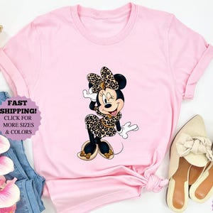 May include: A light pink t-shirt featuring a Minnie Mouse graphic in a leopard print bow, dress, and shoes. The image includes a pair of beige shoes, blue jeans, and a woven purse.