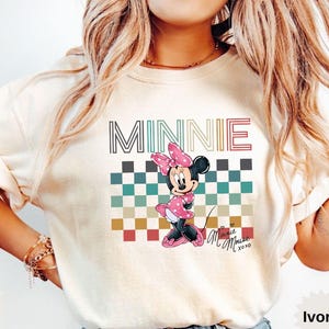 May include: Ivory-colored t-shirt featuring a Minnie Mouse graphic. The design includes the name "MINNIE" in a striped font above a checkered pattern and a cartoon Minnie Mouse wearing a pink dress and bow. The shirt has short sleeves.