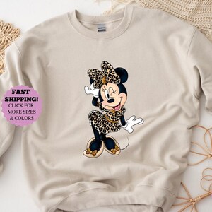 May include: Beige sweatshirt featuring a cartoon Minnie Mouse in a leopard print dress and bow. The image includes text that says "FAST SHIPPING! CLICK FOR MORE SIZES & COLORS".