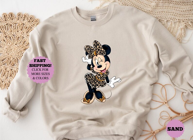 May include: Beige sweatshirt featuring a cartoon Minnie Mouse in a leopard print dress and bow. The image includes text that says "FAST SHIPPING! CLICK FOR MORE SIZES & COLORS".