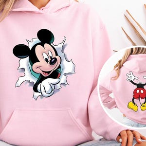 Puede incluir: Sudadera con capucha rosa claro con un gr&aacute;fico de Mickey Mouse. La parte delantera muestra la cara de Mickey saliendo de un agujero rasgado. La parte trasera muestra el cuerpo de Mickey con los brazos levantados en se&ntilde;al de paz. La sudadera tiene un bolsillo delantero y una capucha.