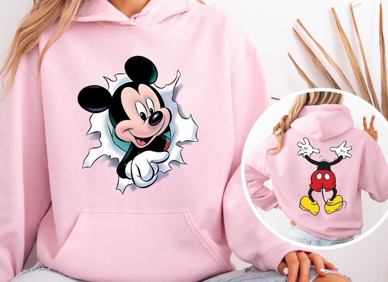 Puede incluir: Sudadera con capucha rosa claro con un gr&aacute;fico de Mickey Mouse. La parte delantera muestra la cara de Mickey saliendo de un agujero rasgado. La parte trasera muestra el cuerpo de Mickey con los brazos levantados en se&ntilde;al de paz. La sudadera tiene un bolsillo delantero y una capucha.