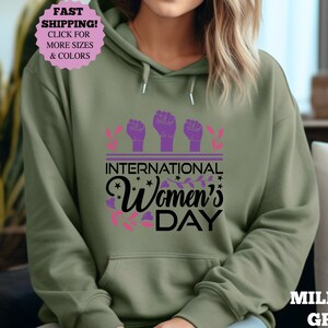 May include: Military green hoodie with a graphic that says "International Women's Day." The design includes three purple fists, floral accents, and the text in black and pink. The hoodie has a front pocket and a drawstring hood.