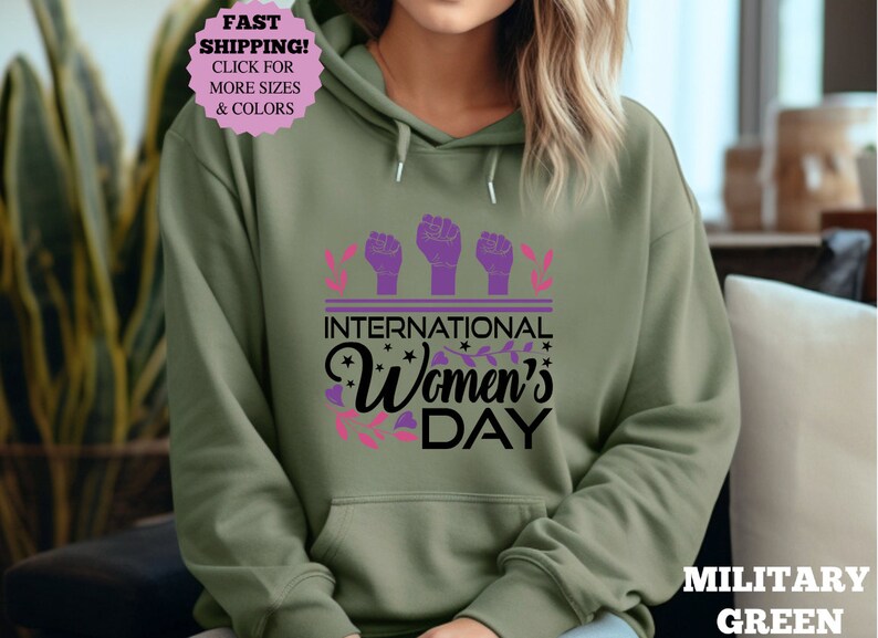 May include: Military green hoodie with a graphic that says "International Women's Day." The design includes three purple fists, floral accents, and the text in black and pink. The hoodie has a front pocket and a drawstring hood.