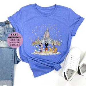 May include: A heather Columbia blue t-shirt featuring a colorful illustration of Disney characters in front of a castle. The shirt is paired with blue jeans and white sneakers. The text "FAST SHIPPING! CLICK FOR MORE SIZES & COLORS" is visible.