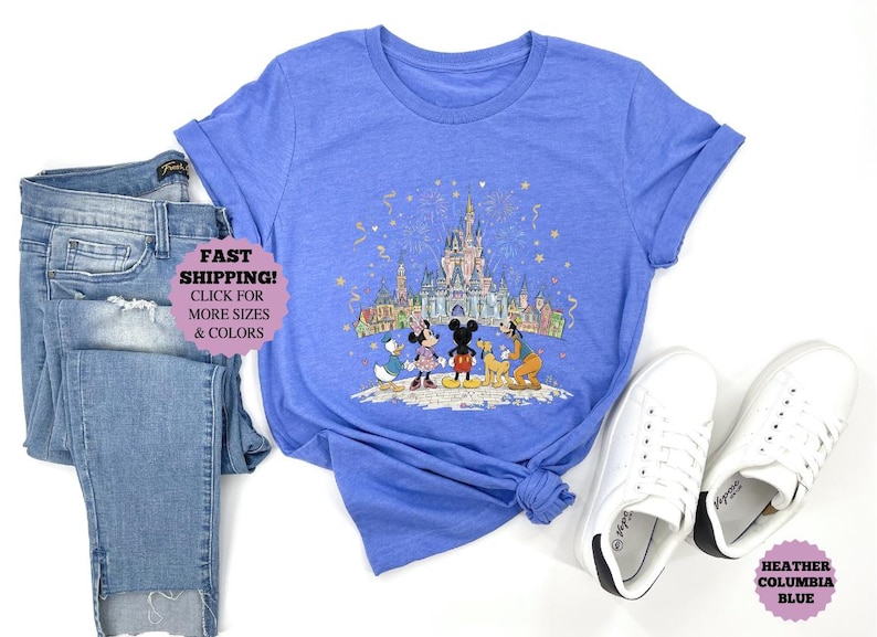 May include: A heather Columbia blue t-shirt featuring a colorful illustration of Disney characters in front of a castle. The shirt is paired with blue jeans and white sneakers. The text "FAST SHIPPING! CLICK FOR MORE SIZES & COLORS" is visible.