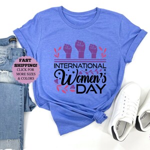 May include: A light blue t-shirt with the text "INTERNATIONAL Women's DAY" in black and purple. The shirt is paired with blue jeans and white sneakers. The shirt has rolled-up sleeves.