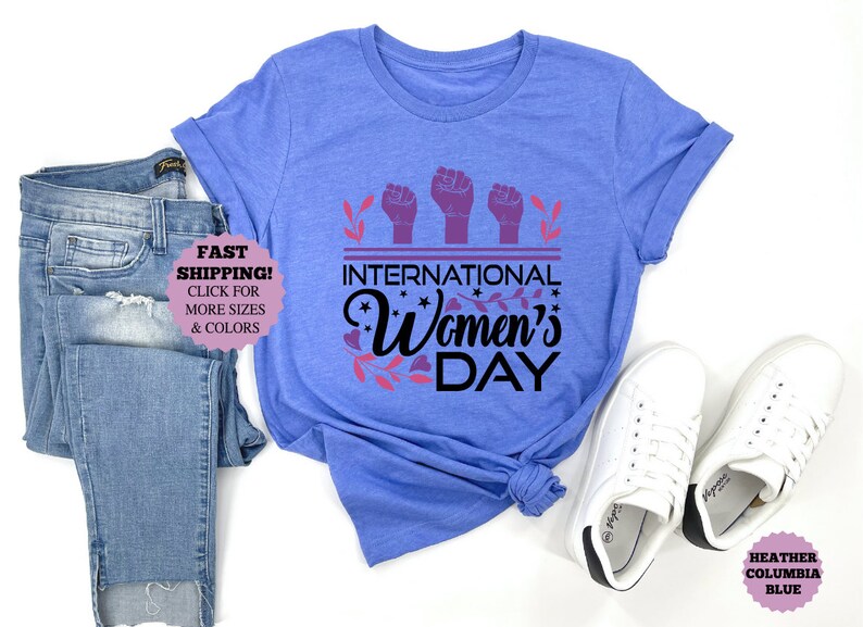 May include: A light blue t-shirt with the text "INTERNATIONAL Women's DAY" in black and purple. The shirt is paired with blue jeans and white sneakers. The shirt has rolled-up sleeves.