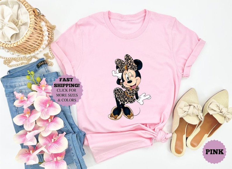 May include: A light pink t-shirt featuring a Minnie Mouse graphic in a leopard print bow, dress, and shoes. The image includes a pair of beige shoes, blue jeans, and a woven purse.