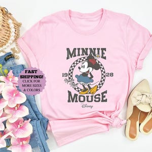 May include: A light pink t-shirt featuring a vintage Minnie Mouse graphic. The design includes the text "MINNIE MOUSE" and "1928". The image also shows blue jeans, a straw bag, and cream-colored shoes.