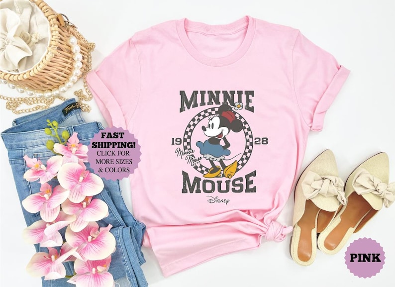 May include: A light pink t-shirt featuring a vintage Minnie Mouse graphic. The design includes the text "MINNIE MOUSE" and "1928". The image also shows blue jeans, a straw bag, and cream-colored shoes.