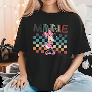 May include: Black t-shirt featuring a graphic of Minnie Mouse in a pink polka-dot dress and bow, standing in front of a checkered pattern. The word "MINNIE" is above the graphic in colorful letters.