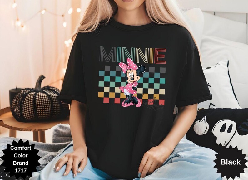 May include: Black t-shirt featuring a graphic of Minnie Mouse in a pink polka-dot dress and bow, standing in front of a checkered pattern. The word "MINNIE" is above the graphic in colorful letters.