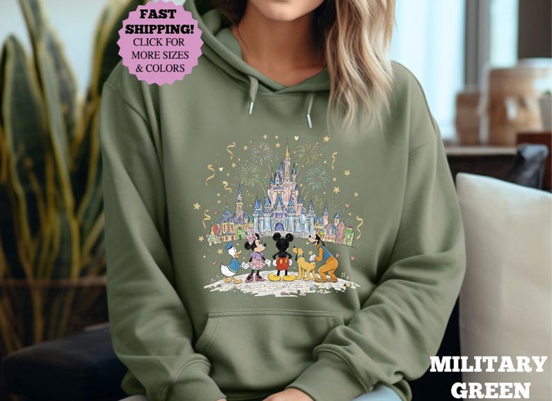 May include: Military green hoodie featuring a colorful graphic of a castle, fireworks, and cartoon characters. The design includes Mickey and Minnie Mouse, Donald Duck, and Goofy. The hoodie has a drawstring hood and a front pocket.