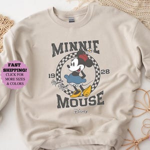 May include: Beige sweatshirt featuring a distressed graphic of Minnie Mouse. The design includes the text "MINNIE MOUSE" and the year "1928". The image also includes the text "CLICK FOR MORE SIZES & COLORS".